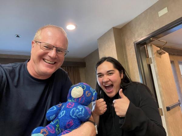 Glen on the left and the Andrea on the right with her thumbs up, are taking a selfie with Peace Bear - a dark blue bear.